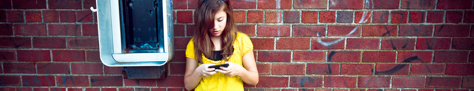 Student in a yellow shirt texts while leaning on a red brick wall